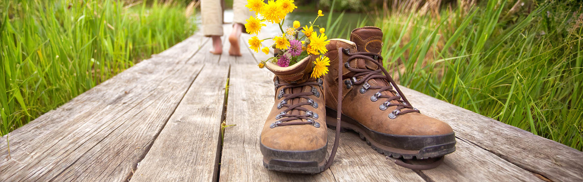 Ansicht eines Schuhpaares mit Blumenstrauß auf einem Ufersteg, im Hintergrund sind Füße
