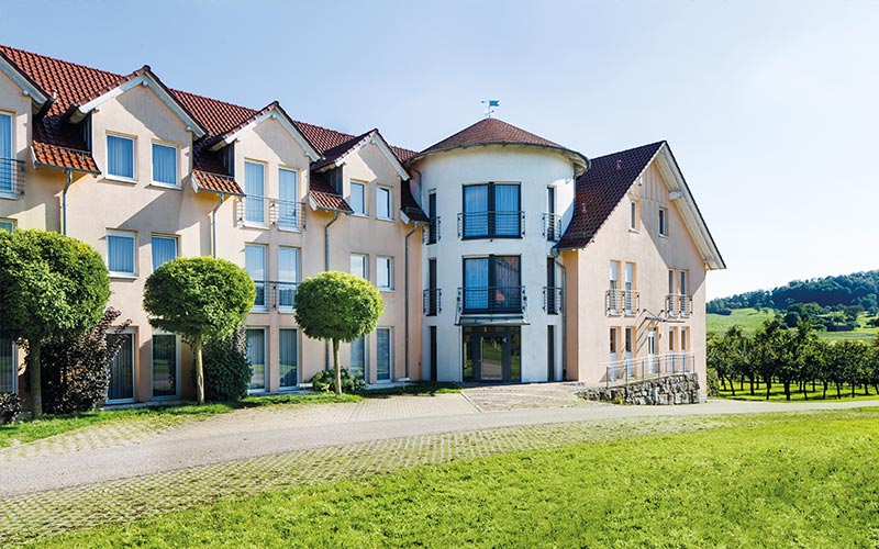 Außenansicht auf das Landhotel Beck und im Hintergrund ist eine Baumplantage und ein Berg